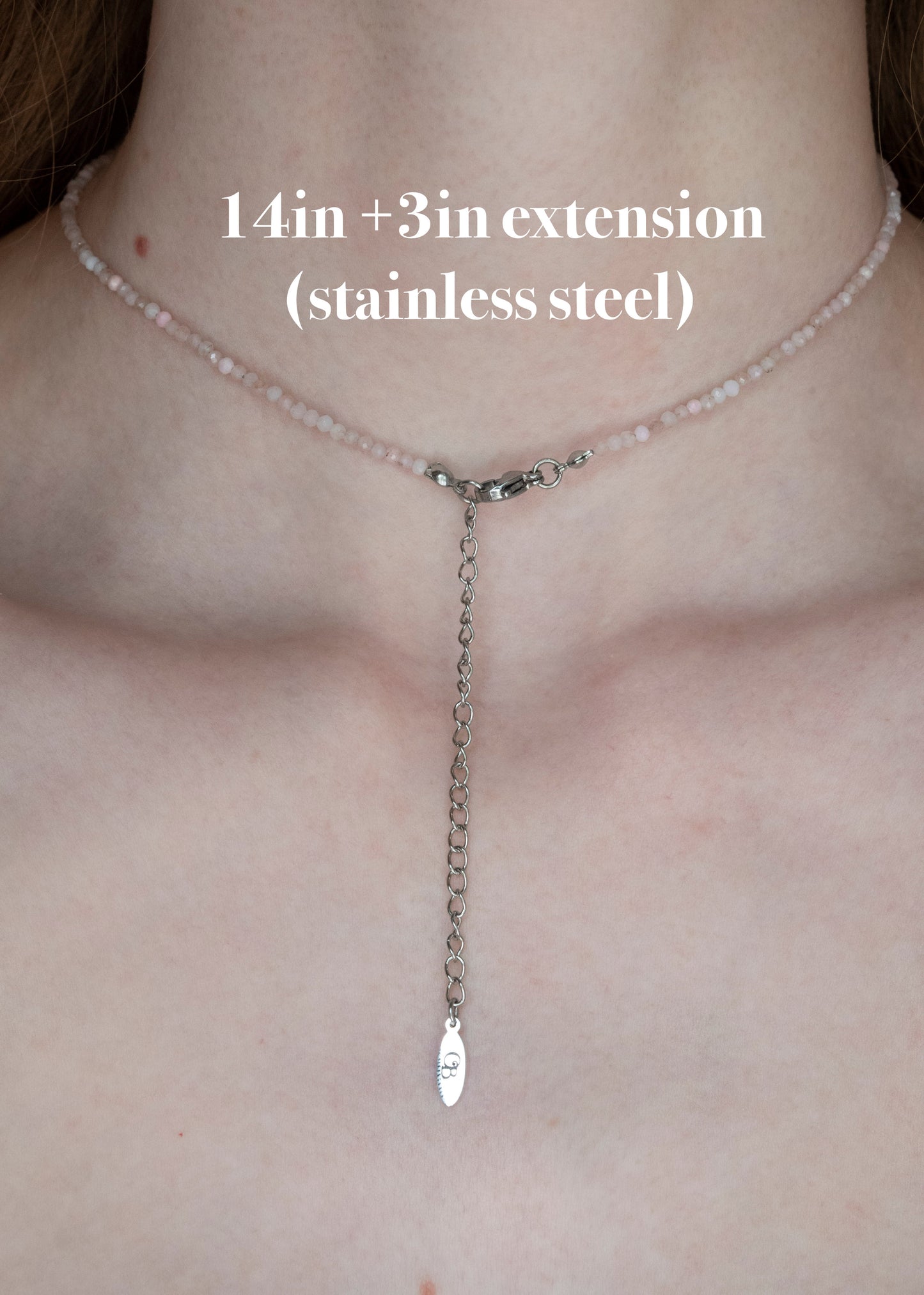 A close-up of a person's neck and upper chest wearing a delicate necklace with a short chain of pale pink beads and a long stainless steel Y-shaped extension ending in a small fish-shaped charm. White text reads '14in +3in extension (stainless steel)'.