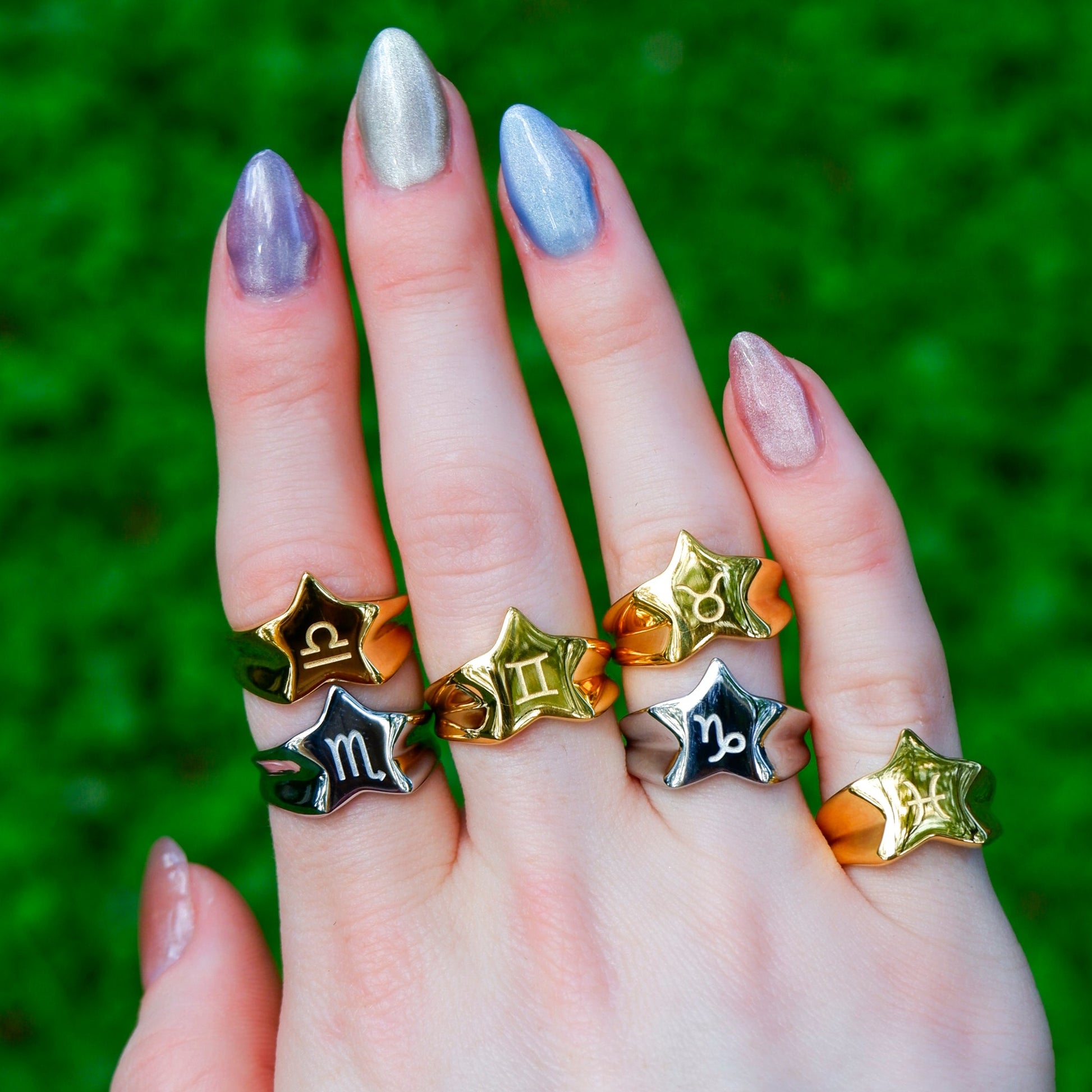 Hand wearing multiple star-shaped rings with different metallic finishes on a green grass background