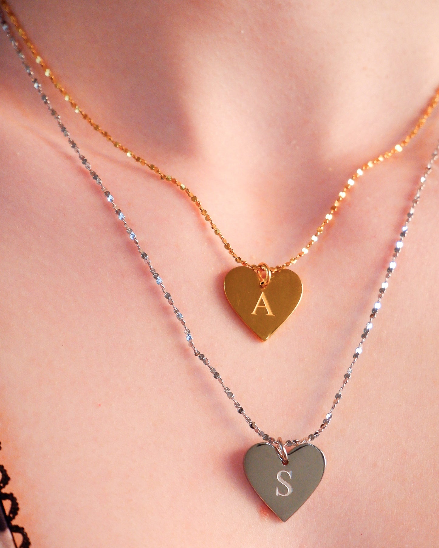 Two heart-shaped pendants on necklaces with initials 'A' and 'S' against a neutral background.