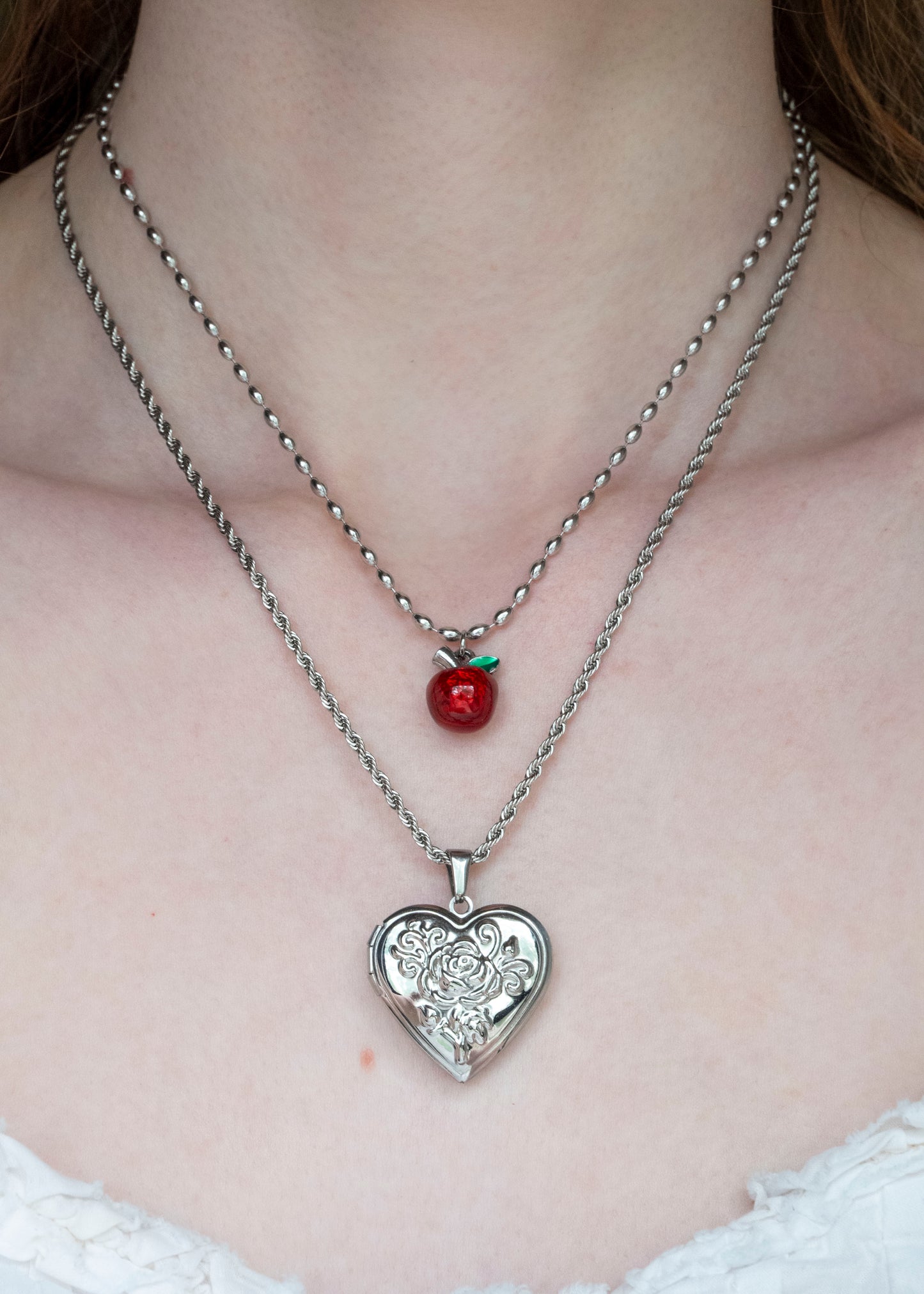 Two necklaces with a heart-shaped pendant and a red apple charm on a person's neck.