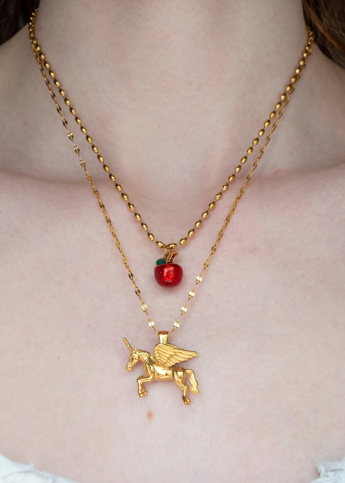 Gold necklace with an alicorn pendant and red apple charm on a person's neck.