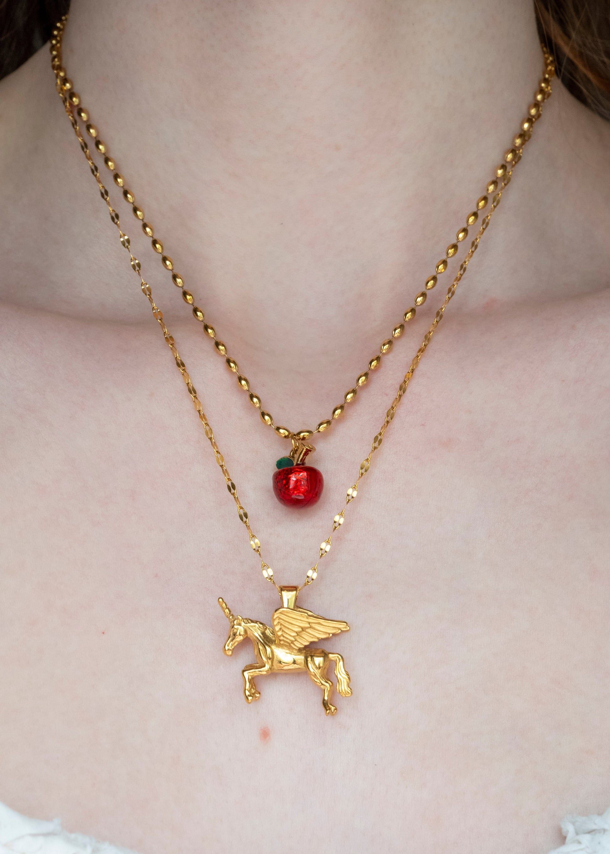Gold necklace with an alicorn pendant and red apple charm on a person's neck.