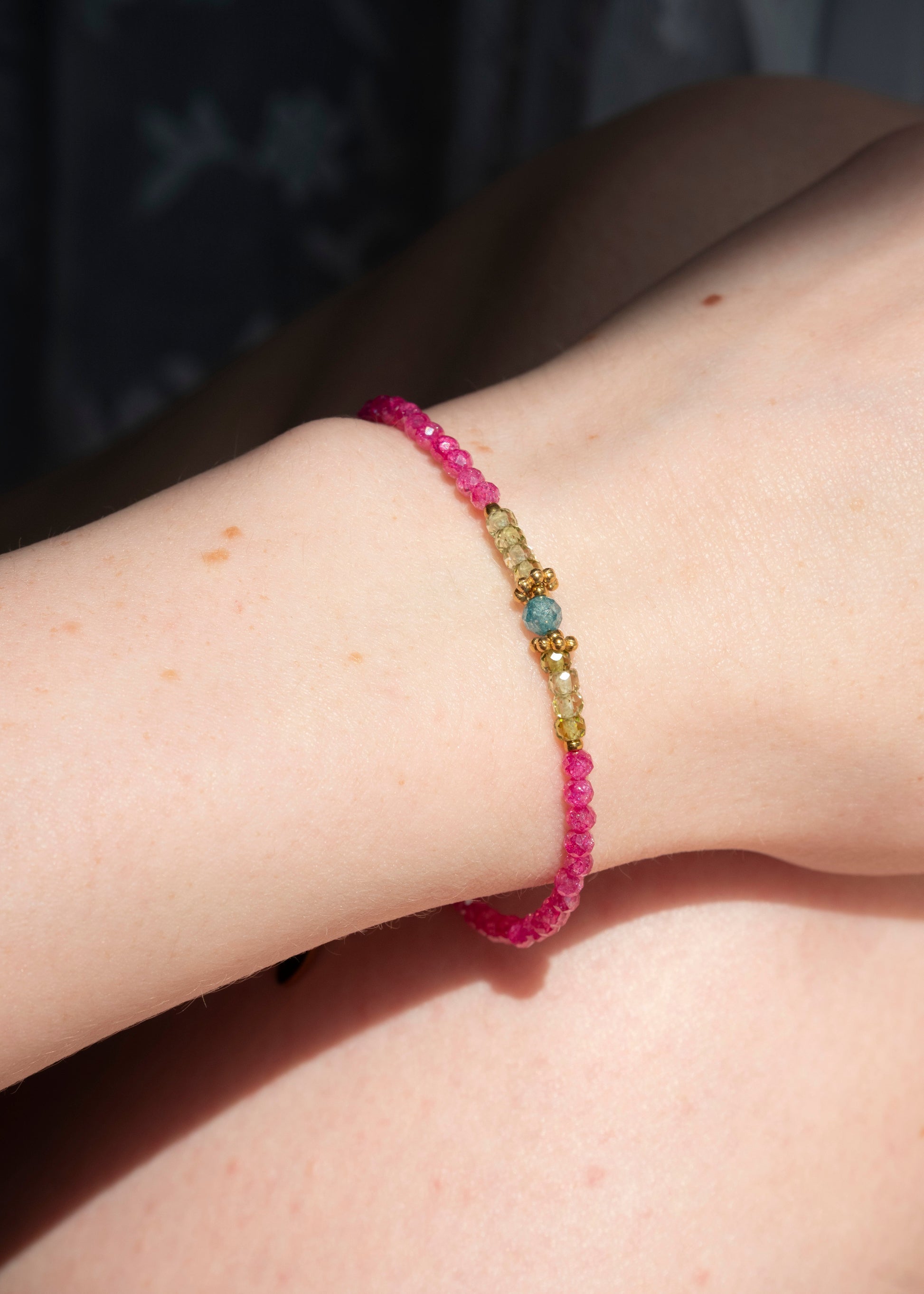 A close-up of a delicate beaded bracelet worn on a person's wrist. The bracelet features a bright pink beaded base with a central gold-toned spacer section containing small yellow, green, and light blue beads.