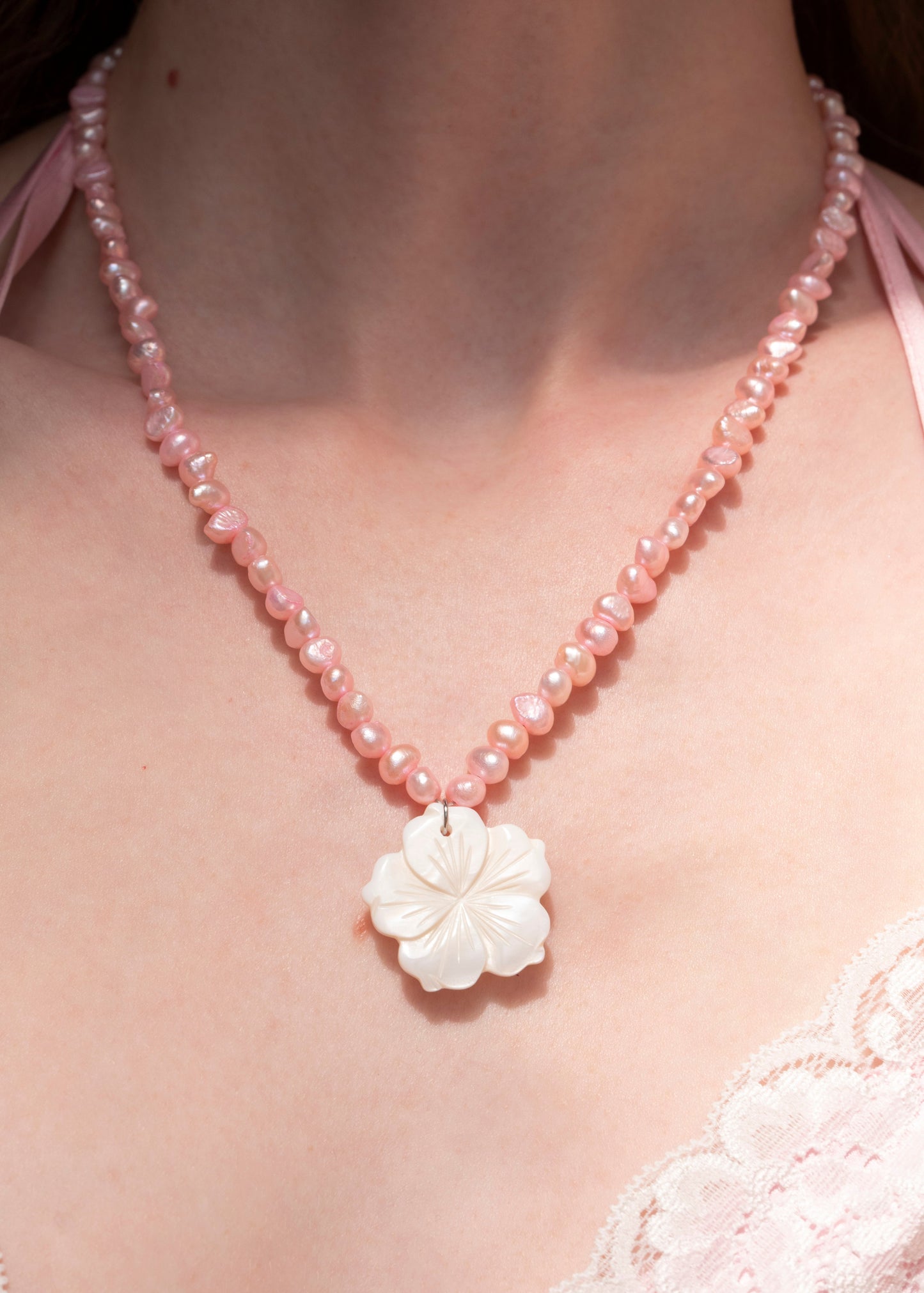 A close-up of a person wearing a pink beaded necklace with a white carved flower pendant, paired with a light pink lace top.