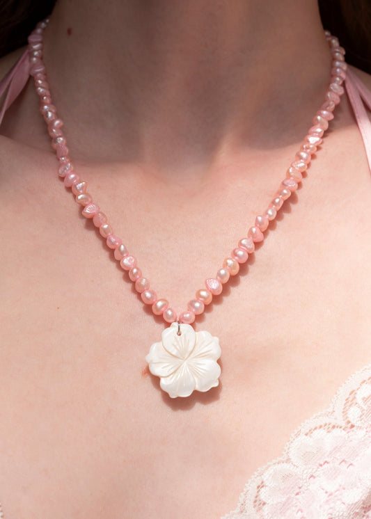 A close-up of a person wearing a pink beaded necklace with a white carved flower pendant, paired with a light pink lace top.