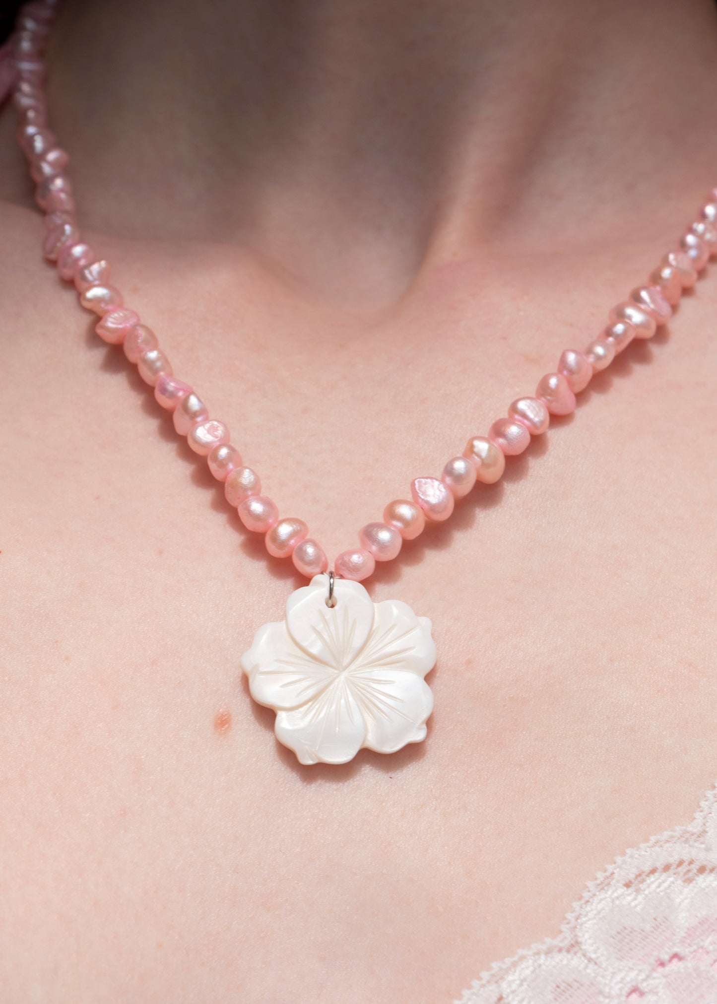 A close-up of a person wearing a pink beaded necklace with a carved white flower pendant, shown against skin and a light pink lace garment.