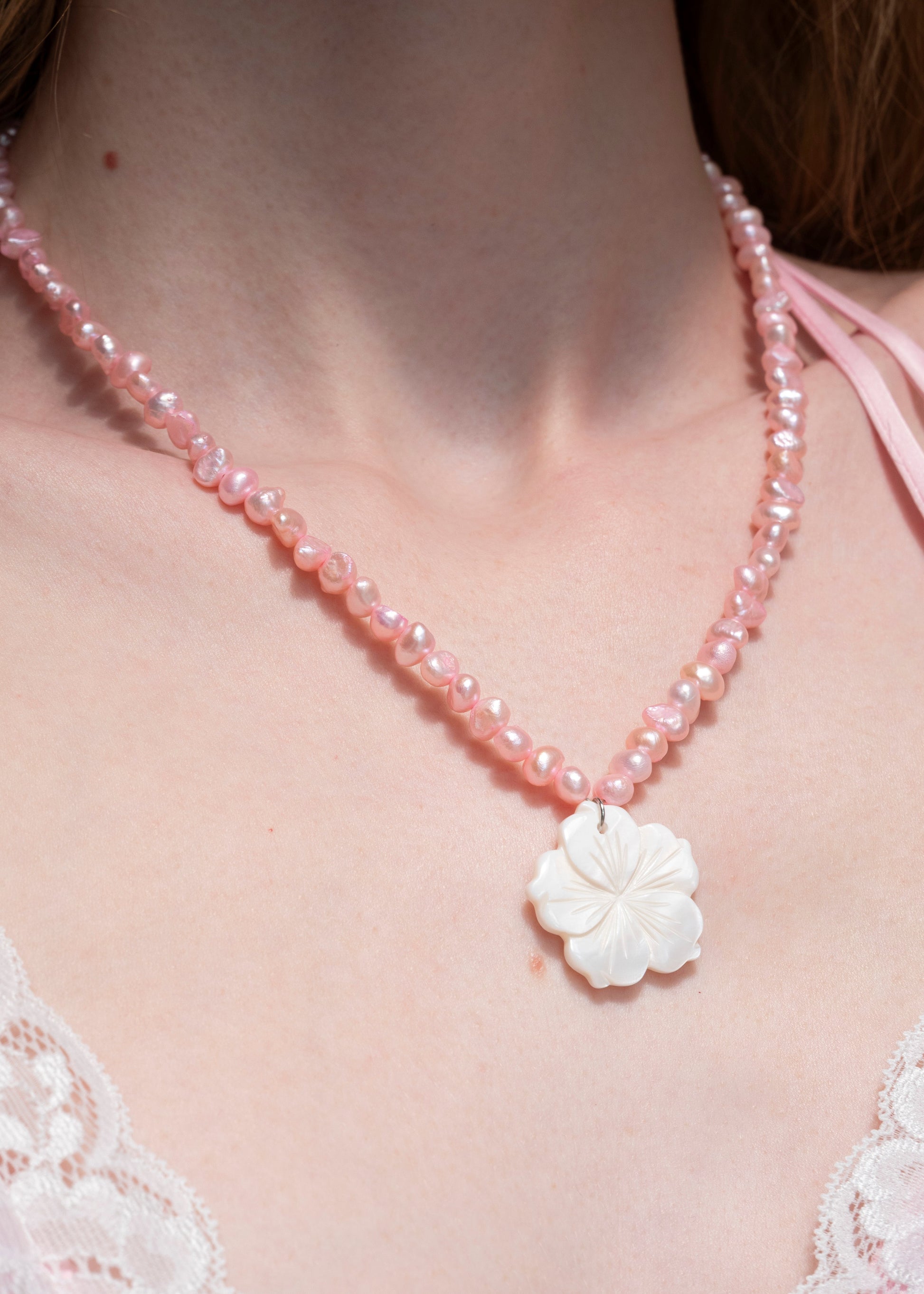 A close-up of a model wearing a pink beaded necklace with a white carved flower pendant, styled with a light pink bralette.