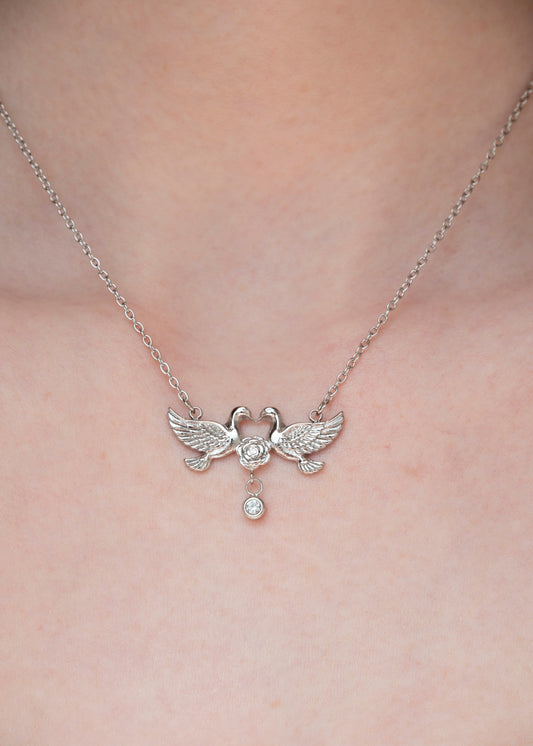 A silver-toned necklace with a pendant shaped like two facing birds holding a small round stone, worn on a person's neck against a light skin background.