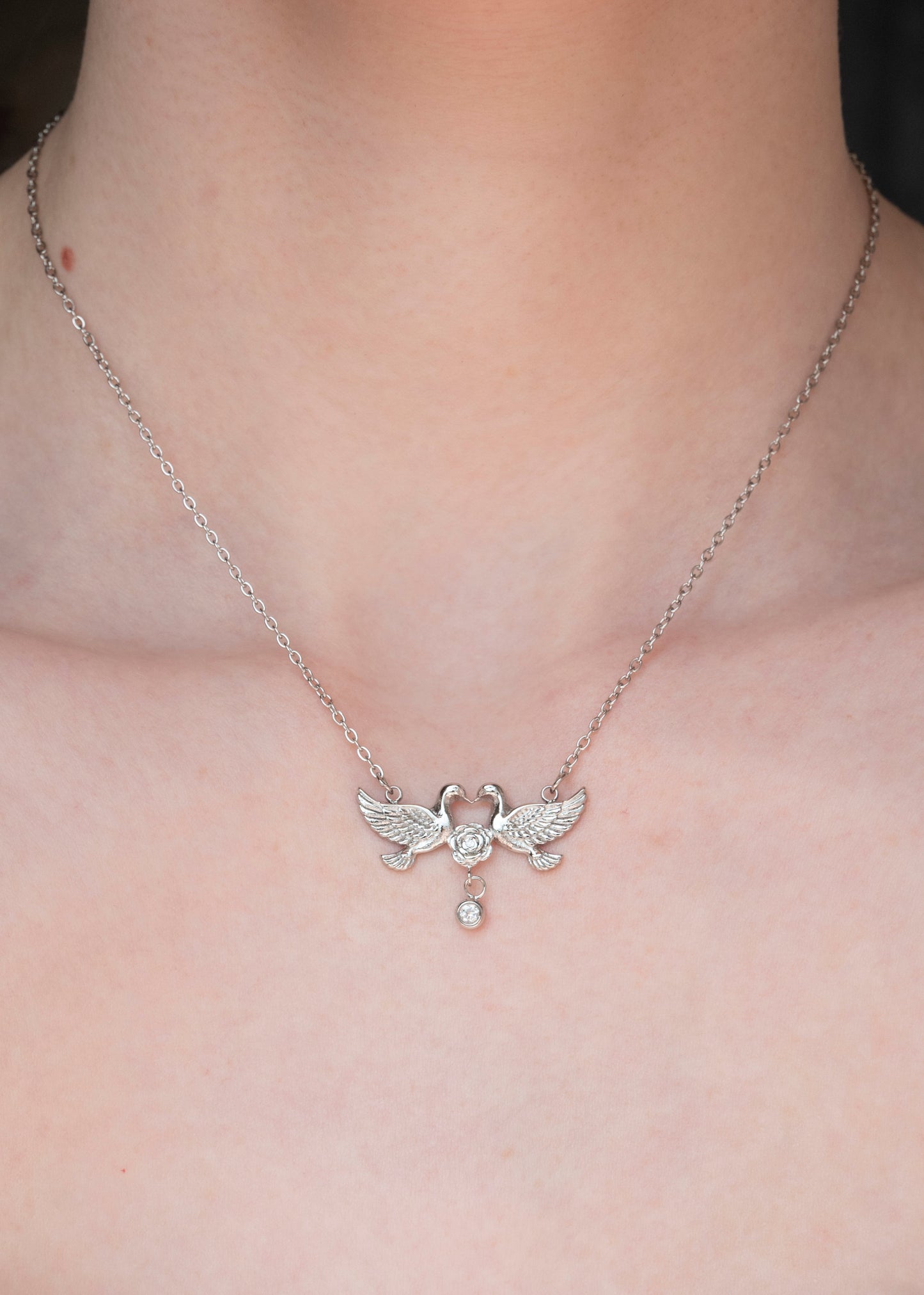A close-up of a person's neck and upper chest wearing a delicate silver chain necklace with a winged pendant and a small dangling pearl.