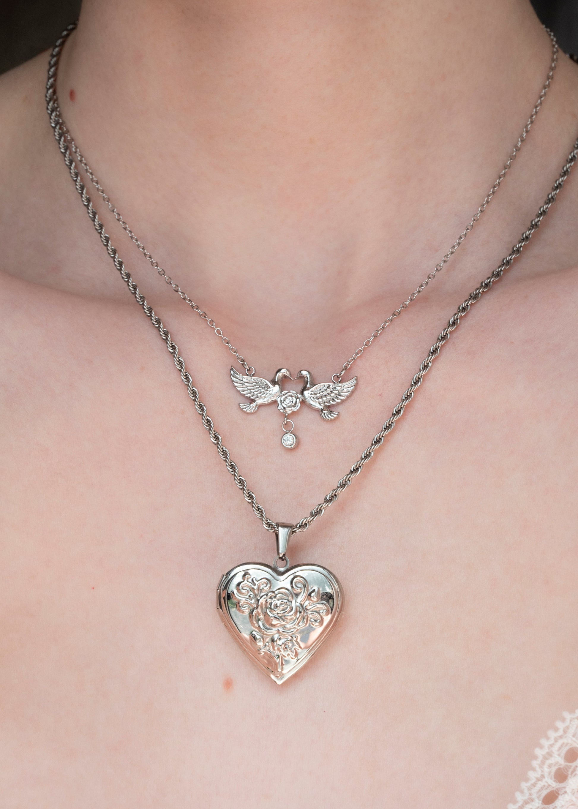 A close-up of a person's neck and chest wearing two layered silver necklaces. The shorter chain features a small winged cherub pendant with a dangling pearl-like bead, while the longer chain holds a larger heart-shaped locket pendant with an ornate floral filigree design.