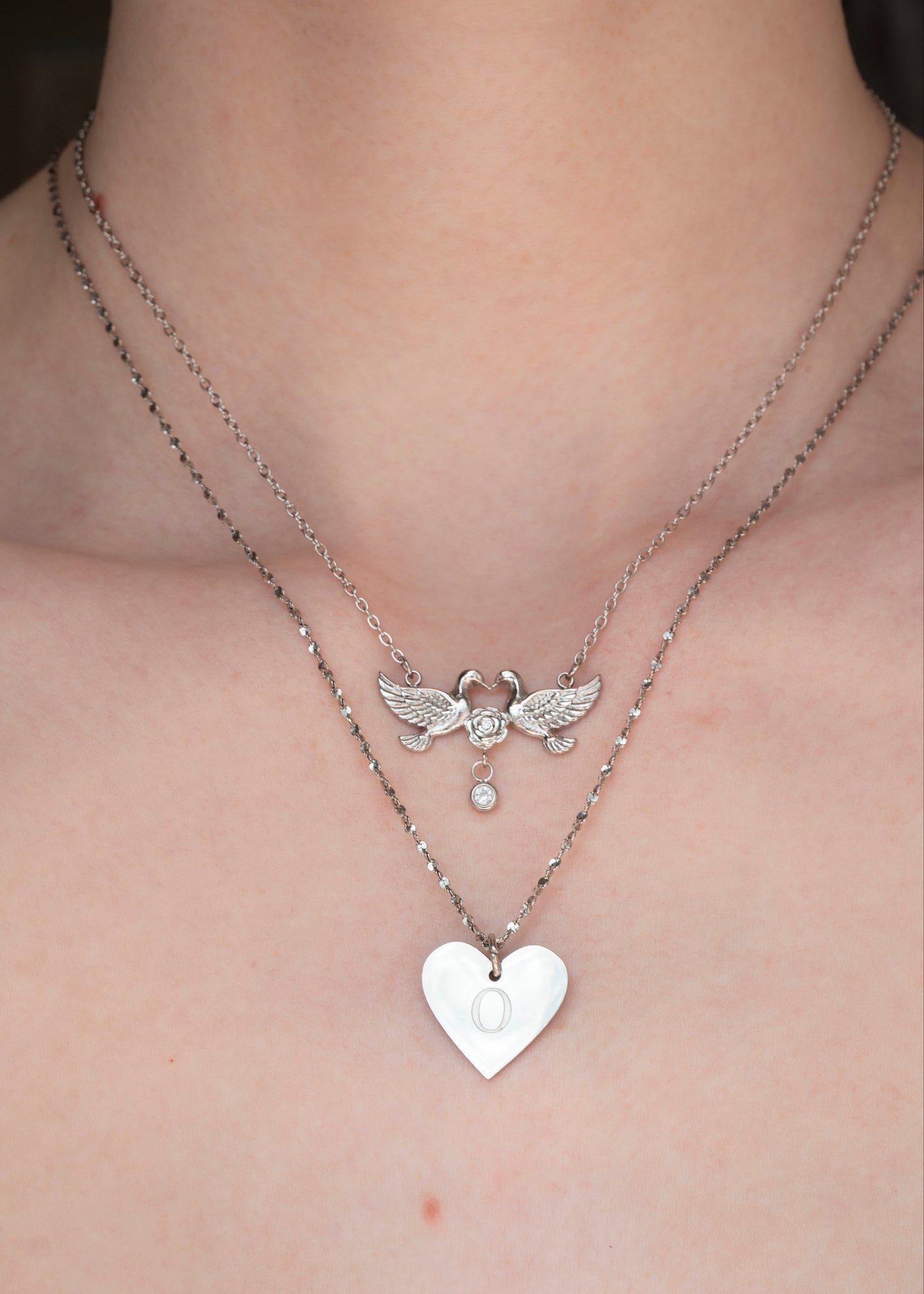 A close-up of a person's neck and upper chest wearing two layered silver necklaces. The shorter chain features a winged angel pendant with a small dangling pearl-like bead, while the longer chain holds a polished silver heart pendant.
