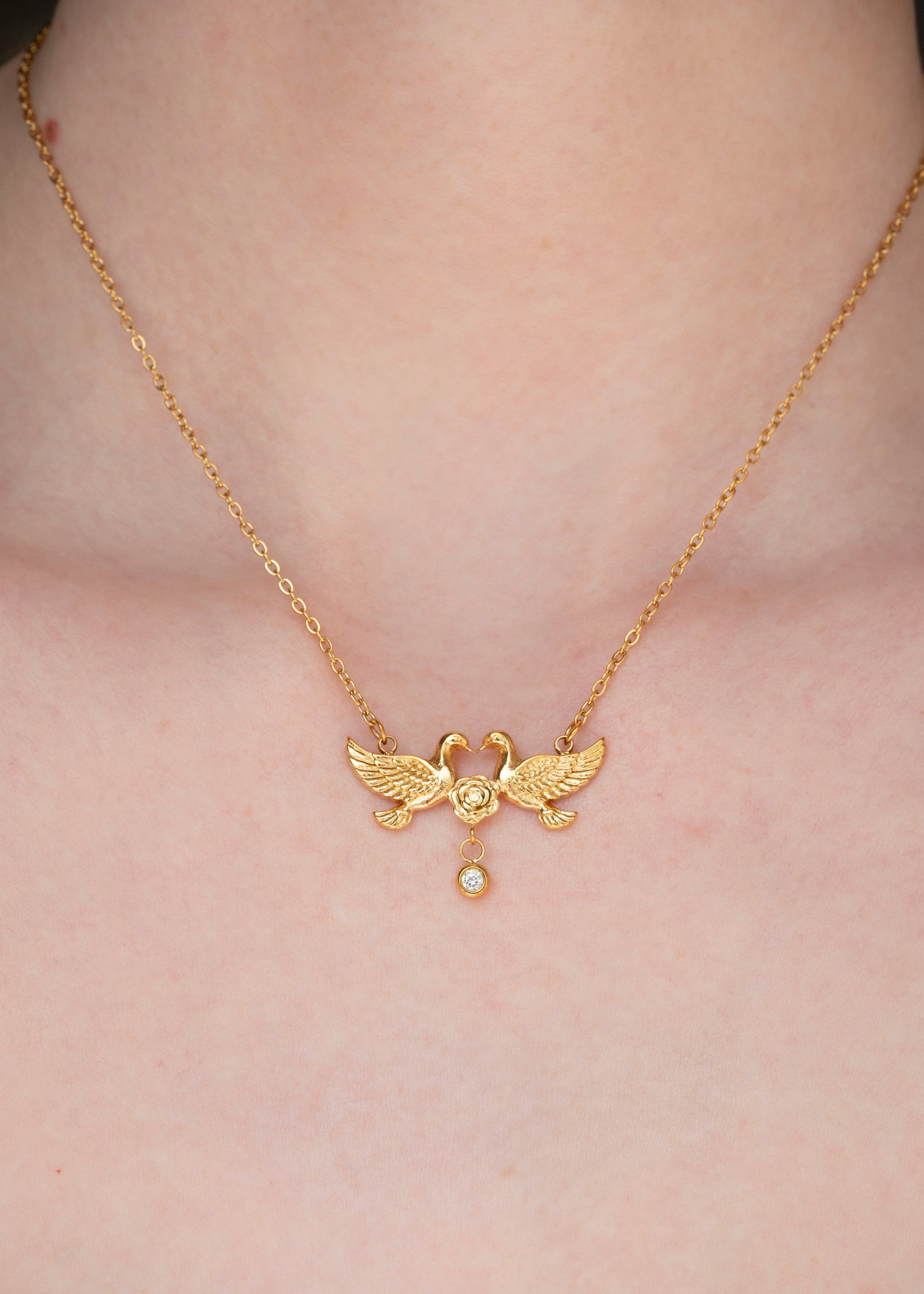 A close-up of a person's neck and chest wearing a delicate gold chain necklace with a swan pendant and a small dangling pearl.