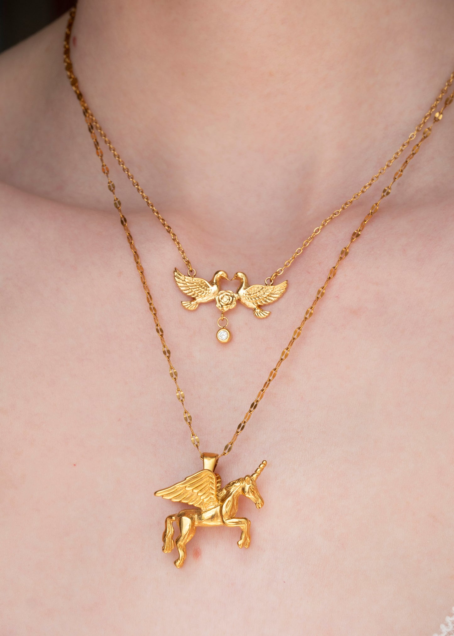 A close-up of a person's neck and chest wearing two layered gold necklaces. The shorter chain features a winged cherub pendant, and the longer chain features a winged unicorn pendant.