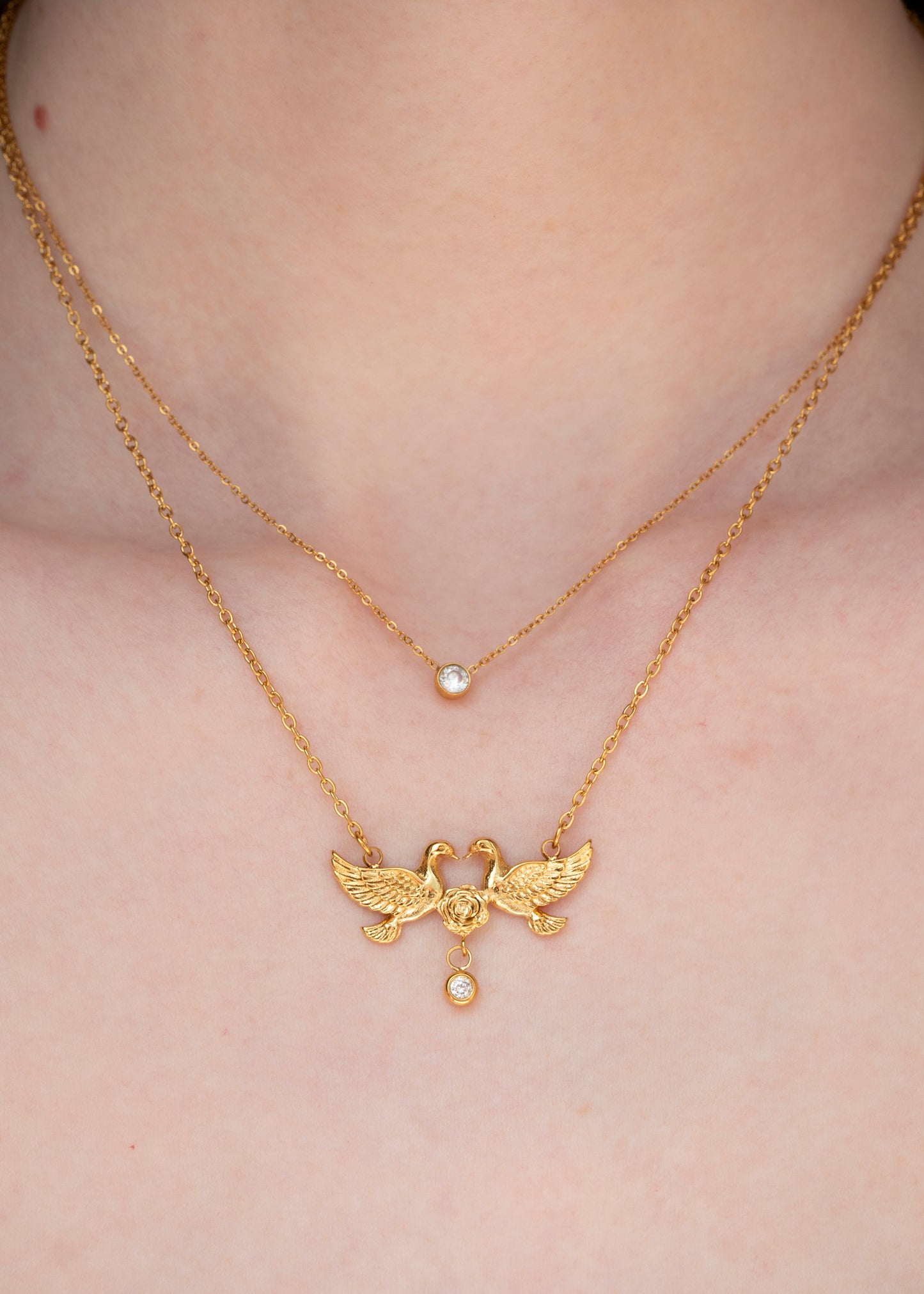 A close-up of a person's neck and chest wearing two layered gold necklaces. The shorter chain has a small round pearl-like bead, while the longer chain features a detailed gold pendant with two birds facing each other and a small hanging pearl bead.