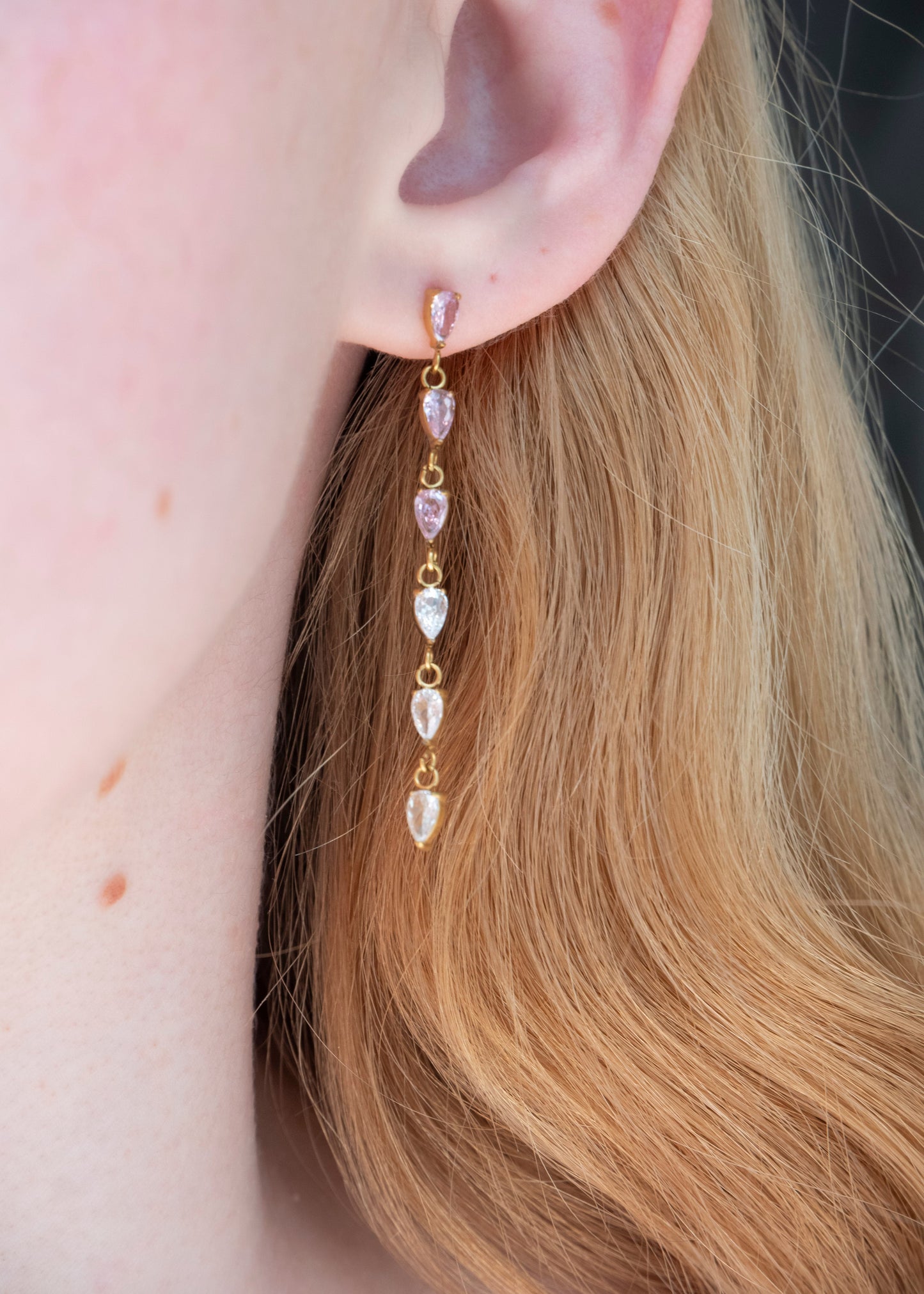 A close-up of a model's ear wearing a gold drop earring with alternating clear teardrop gemstones and light pink teardrop gemstones.