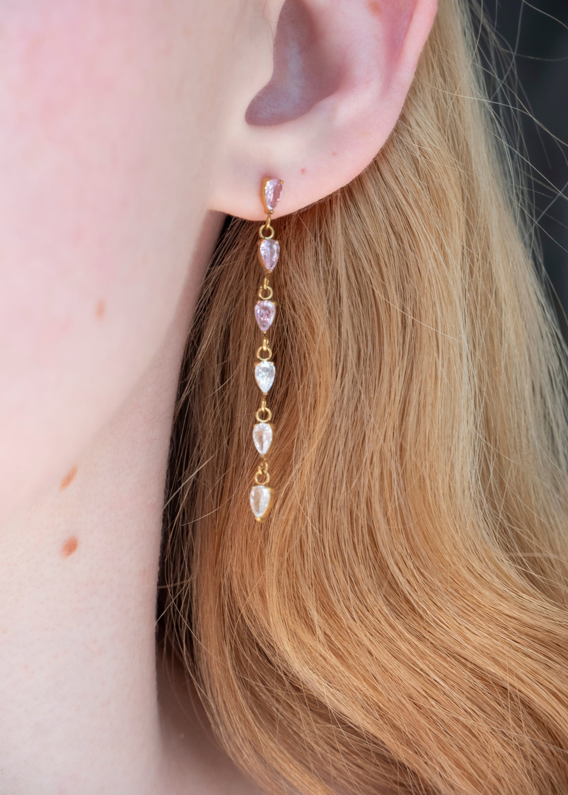 A close-up of a model's ear wearing a gold drop earring with alternating clear teardrop gemstones and light pink teardrop gemstones.
