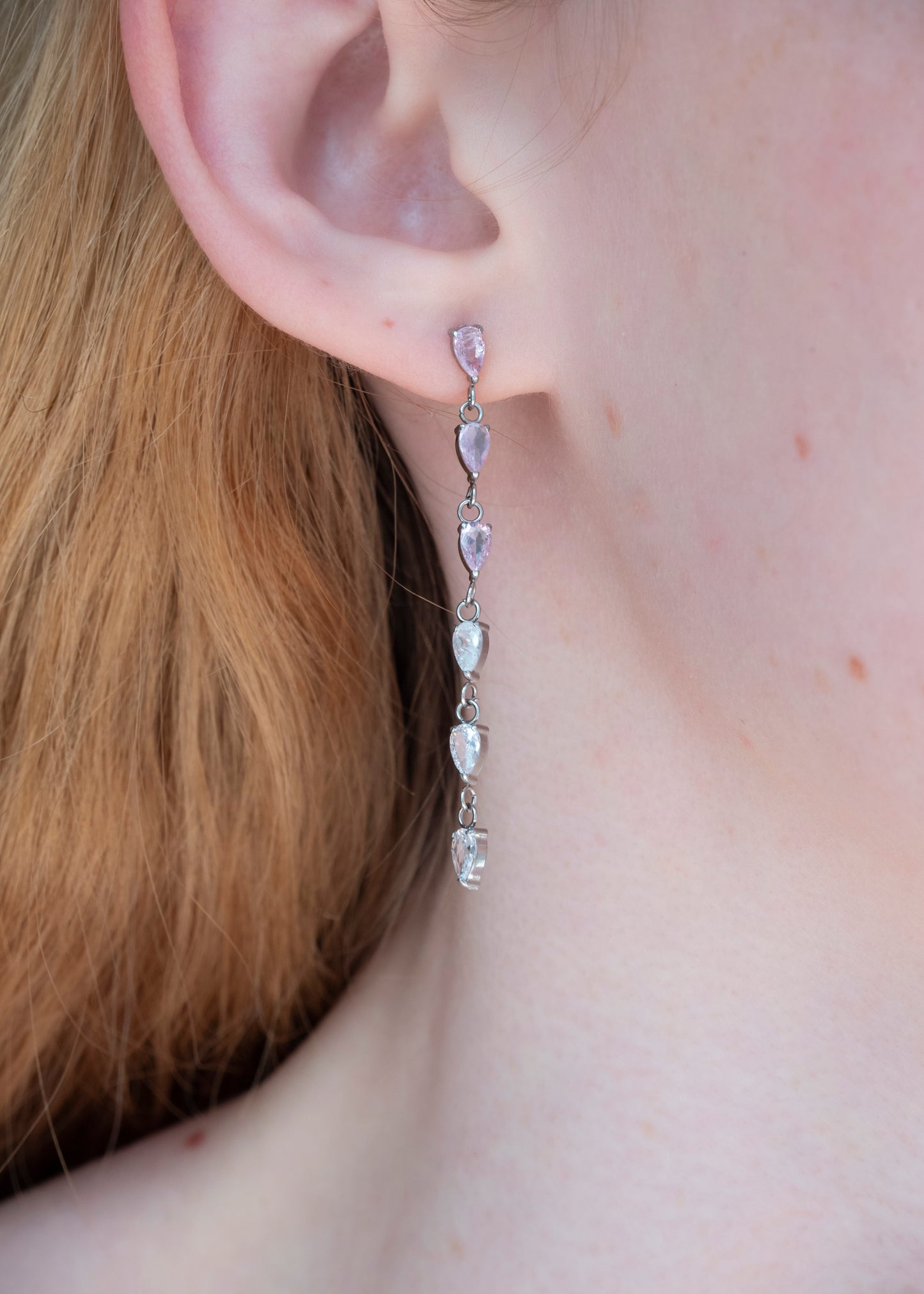 A close-up of a person's ear wearing a long dangling earring with five clear teardrop-shaped stones set in silver-toned metal.