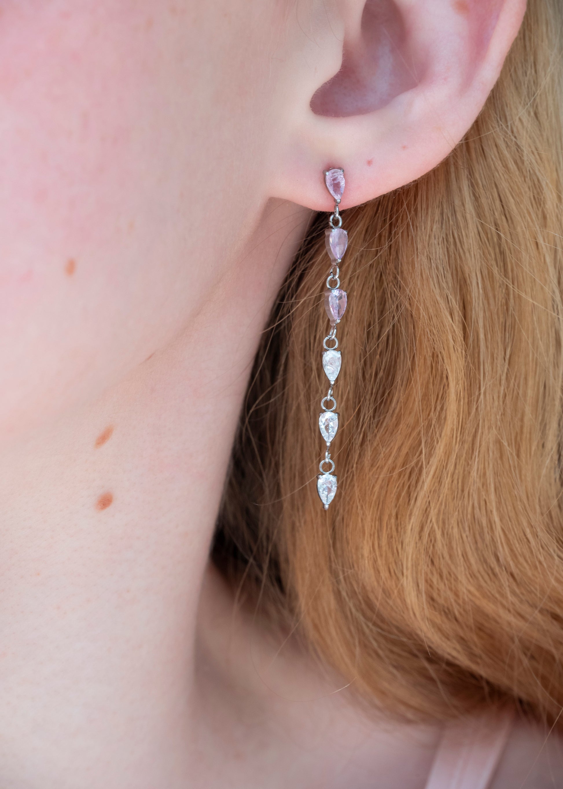 A close-up of a person's ear wearing a dangling earring with small pink faceted beads and silver-toned teardrop charms.