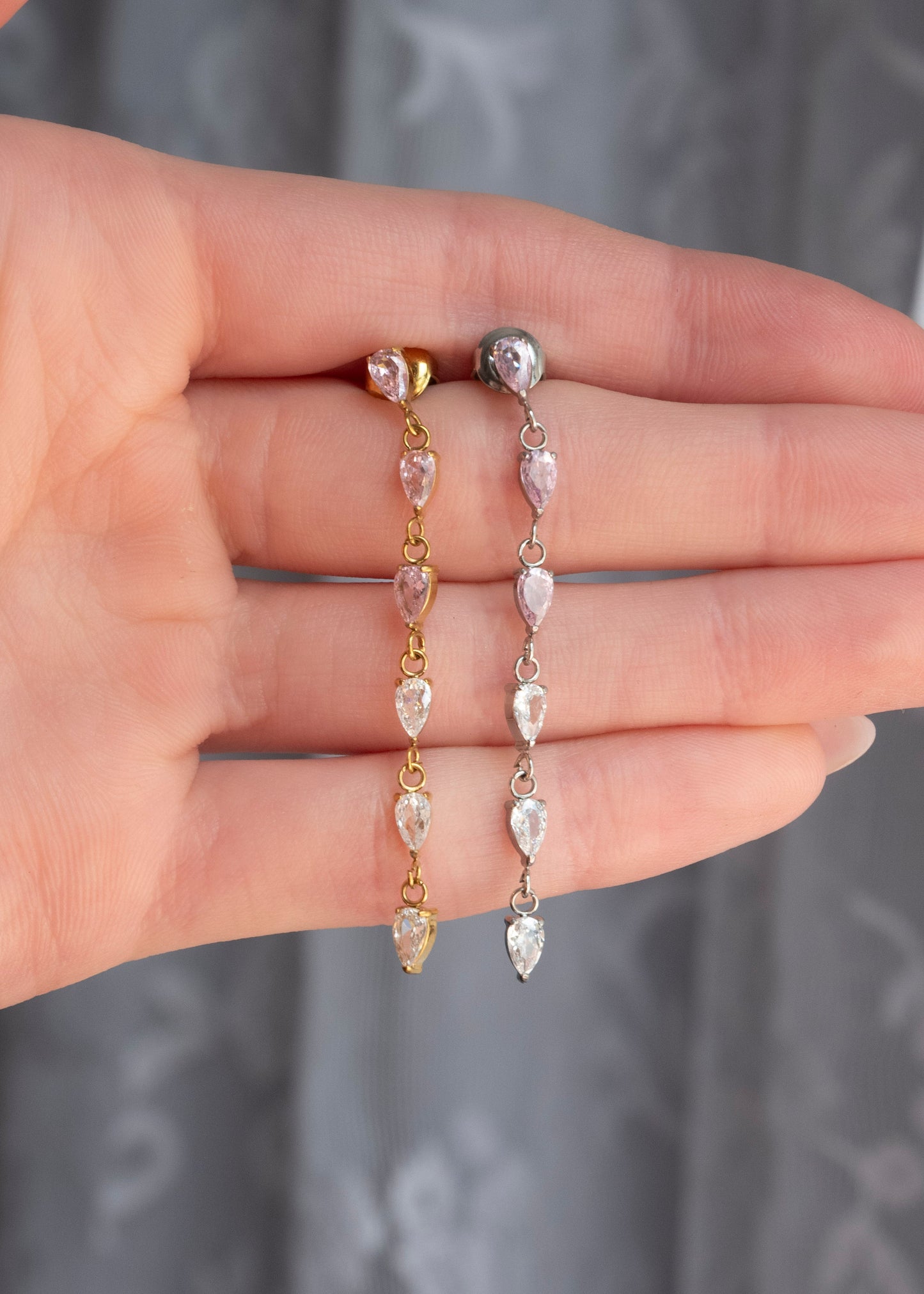 A close-up of a hand holding two long drop earrings with clear teardrop-shaped stones and gold-toned settings, against a gray patterned curtain background.