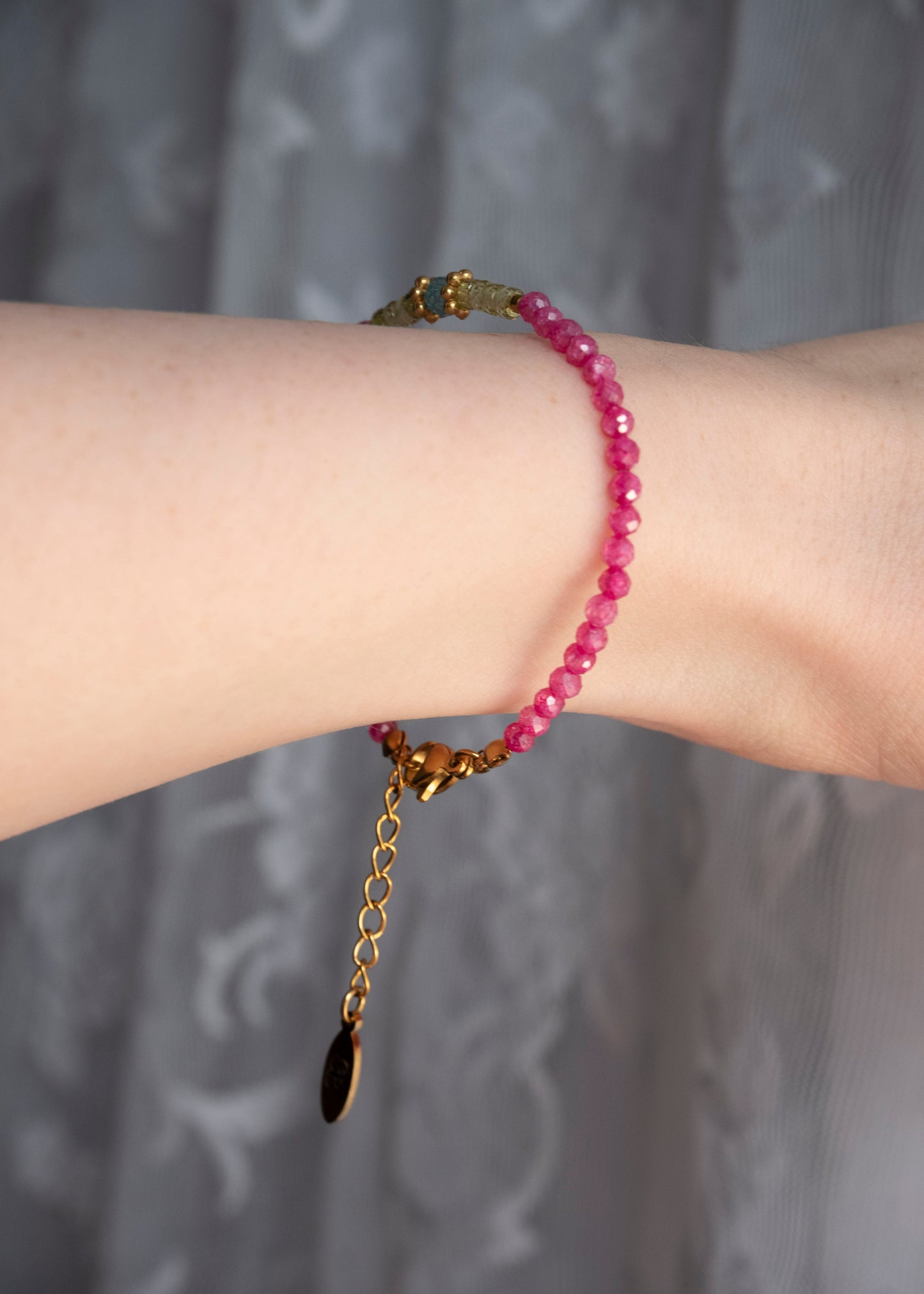 A close-up of a person's wrist wearing a beaded bracelet made of faceted pink beads with gold-toned metal hardware, including a clasp and extender chain with a dark teardrop charm, against a soft gray patterned curtain background.