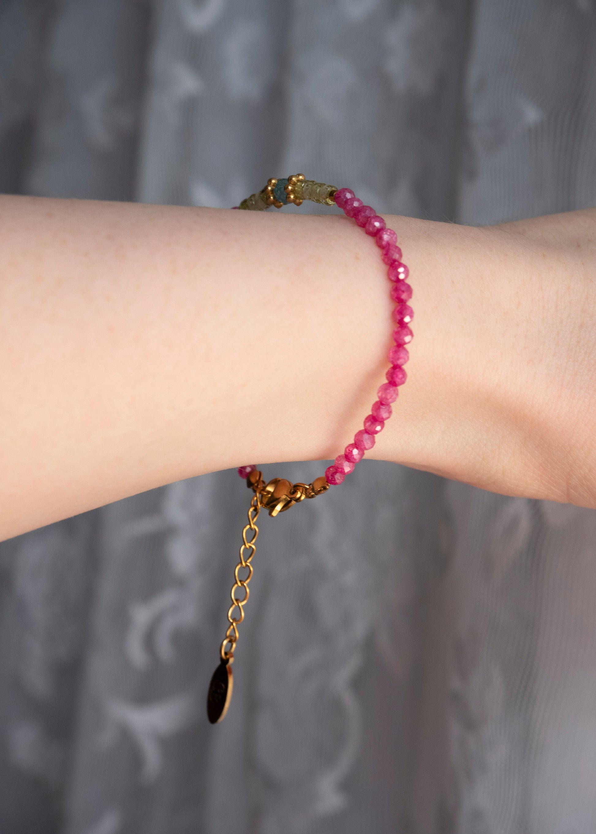 A close-up of a person's wrist wearing a beaded bracelet made of faceted pink beads with gold-toned metal hardware, including a clasp and extender chain with a dark teardrop charm, against a soft gray patterned curtain background.
