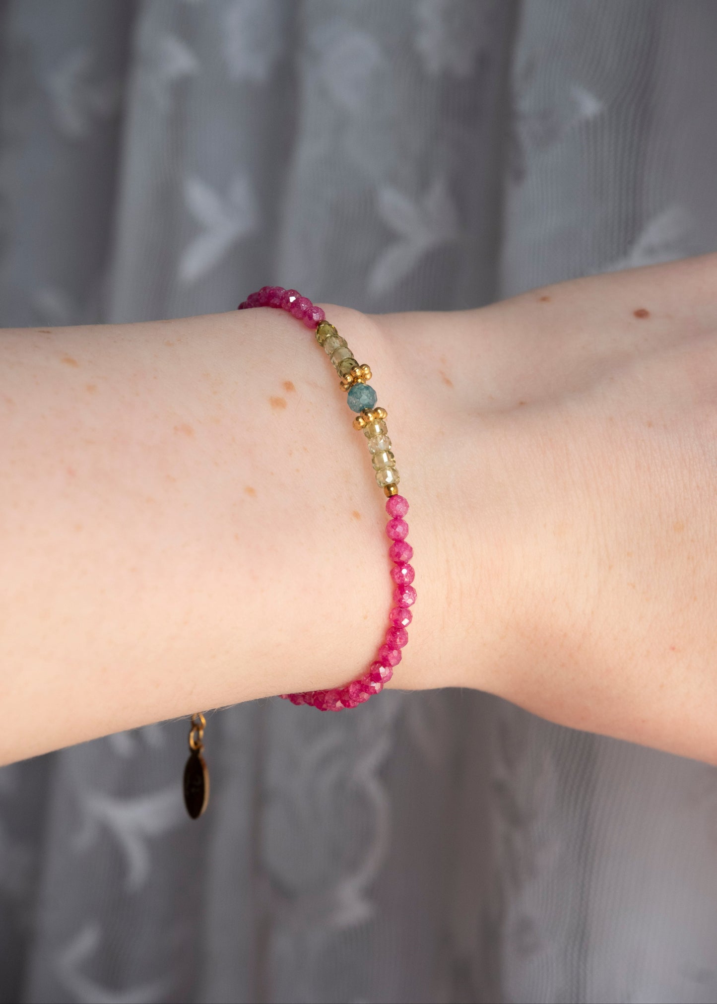 A close-up of a person's wrist wearing a beaded bracelet with pink faceted beads, a small gold spacer, and a light green stone accent, set against a gray patterned curtain background.