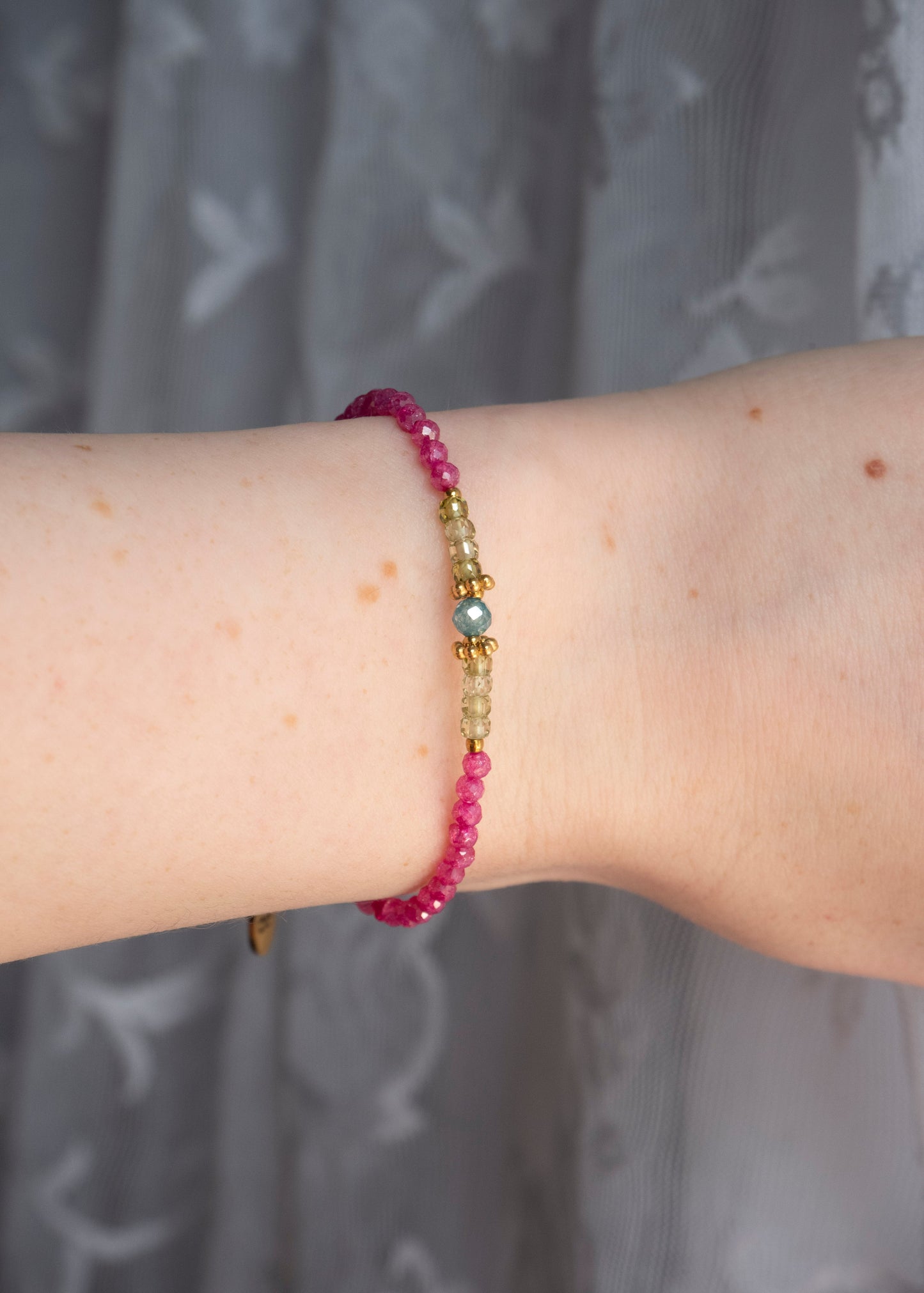 A close-up of a person's wrist wearing a beaded bracelet with pink faceted beads, gold spacer beads, and a central iridescent blue-green bead, set against a soft gray patterned curtain background.
