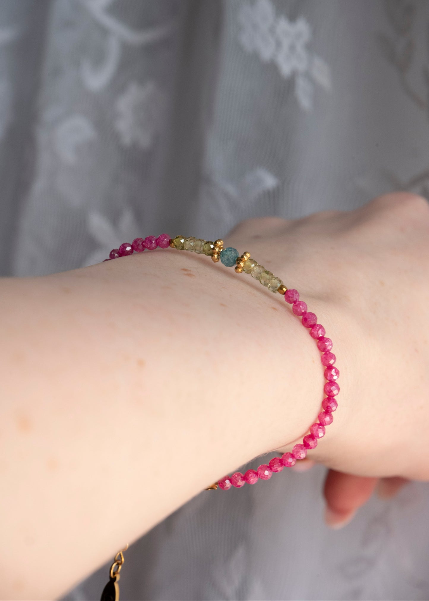 A close-up of a person's wrist wearing a beaded bracelet with bright pink faceted beads, small gold-toned spacer details, and a central round blue-green stone, set against a soft gray patterned fabric background.