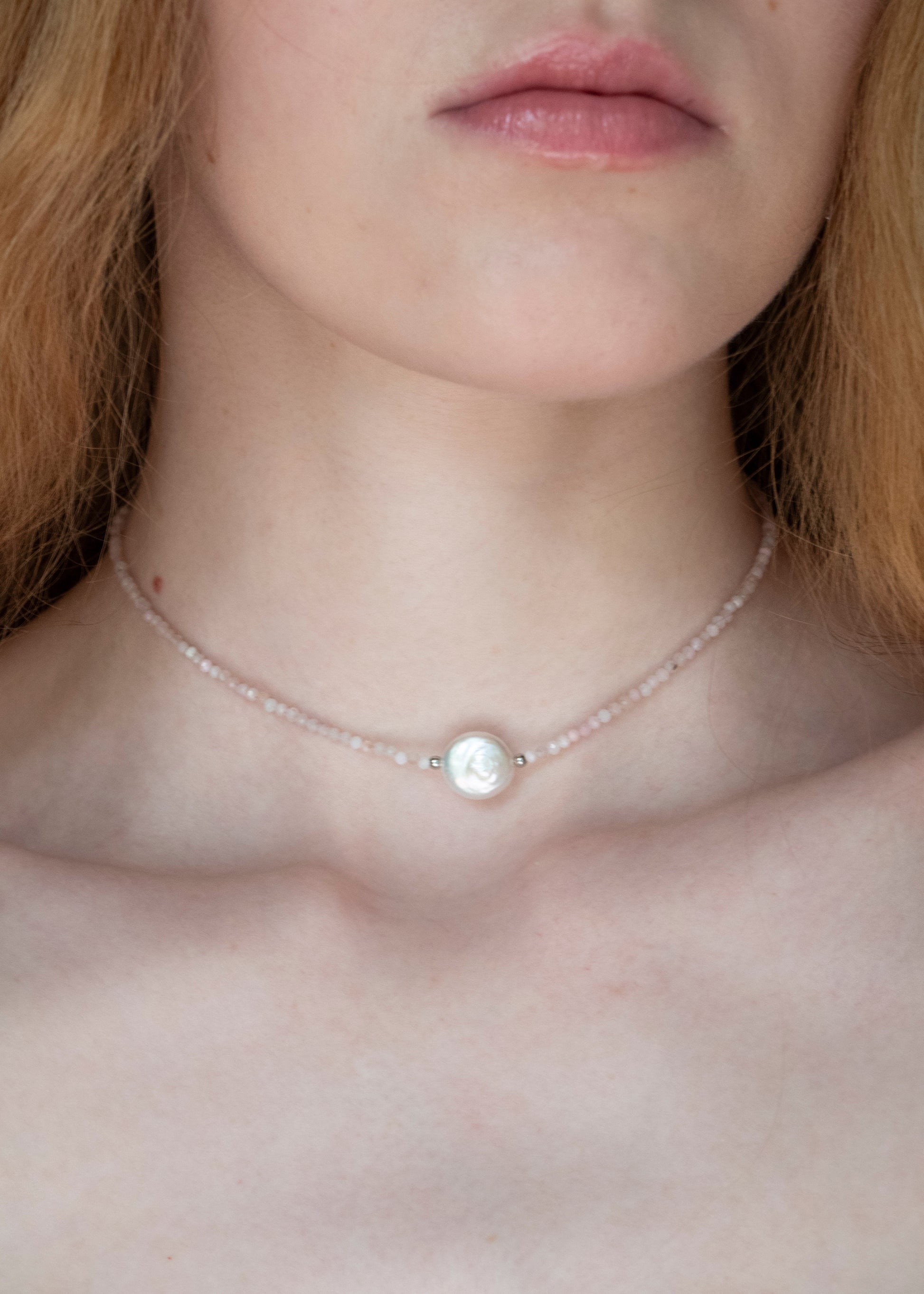 A close-up of a model wearing a delicate choker necklace made of small pale pink beads with a central white pearl-like bead.