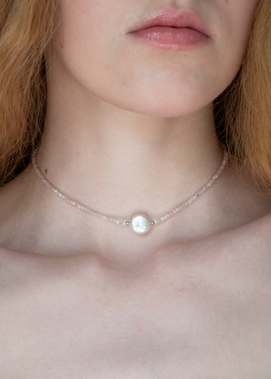 A close-up of a model wearing a delicate choker necklace made of small pale pink beads with a central white pearl-like bead.