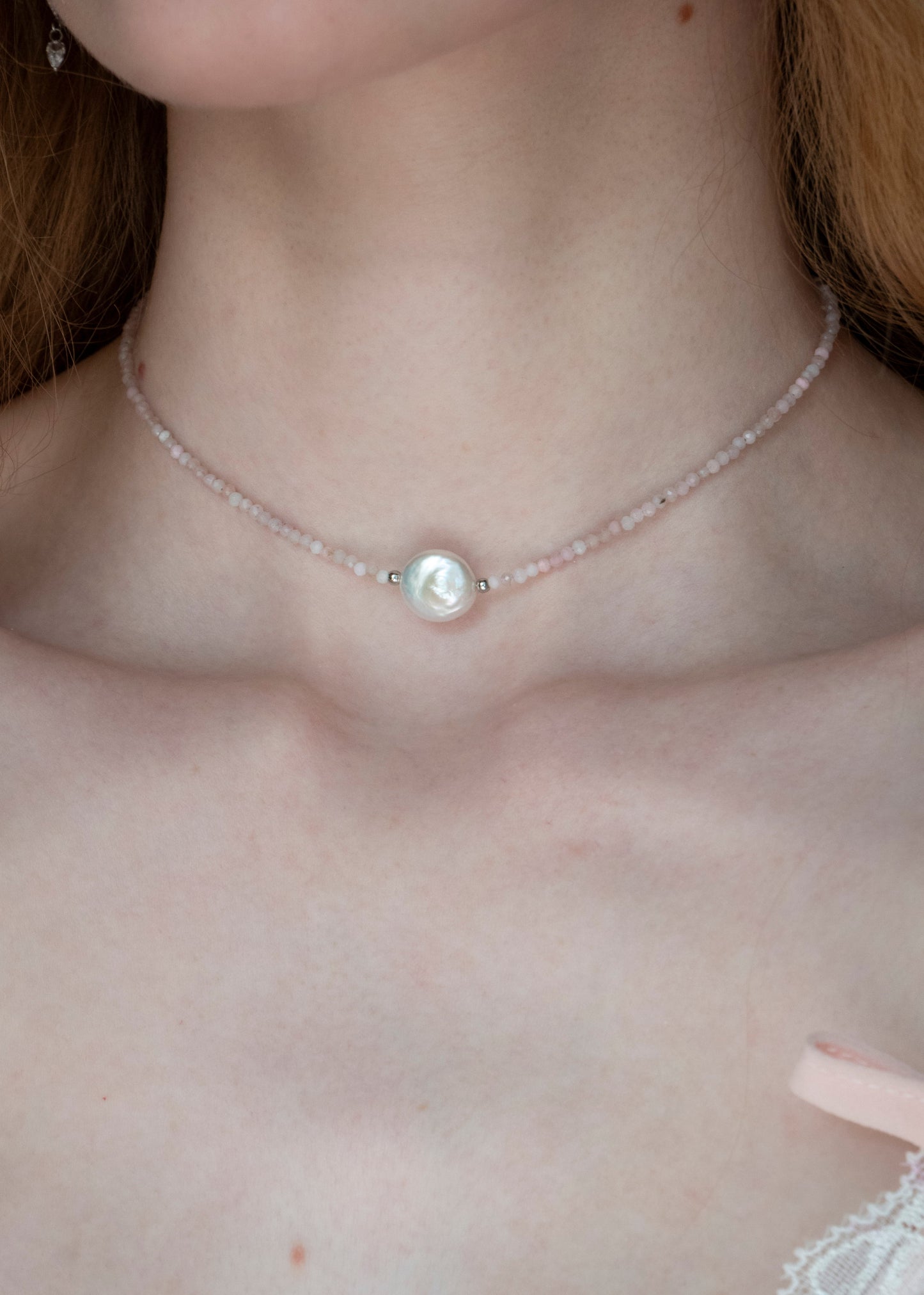 A close-up of a delicate necklace worn on a person's neck, featuring a central white pearl-like bead with a silver-toned setting and a thin beaded chain in a pale pink tone.