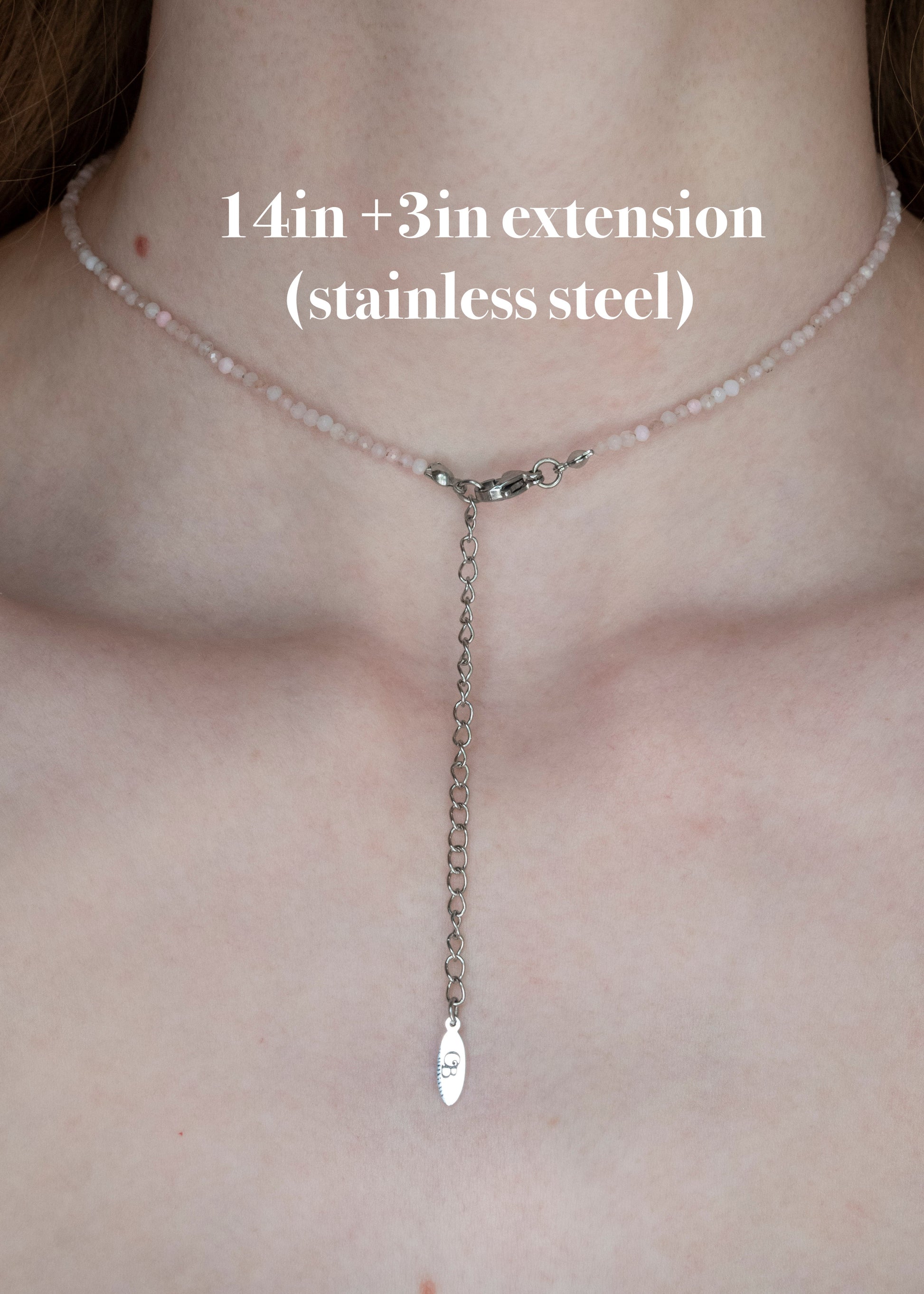 A close-up of a person's neck and upper chest wearing a delicate necklace with a short chain of pale pink beads and a long stainless steel Y-shaped extension ending in a small fish-shaped charm. White text reads '14in +3in extension (stainless steel)'.