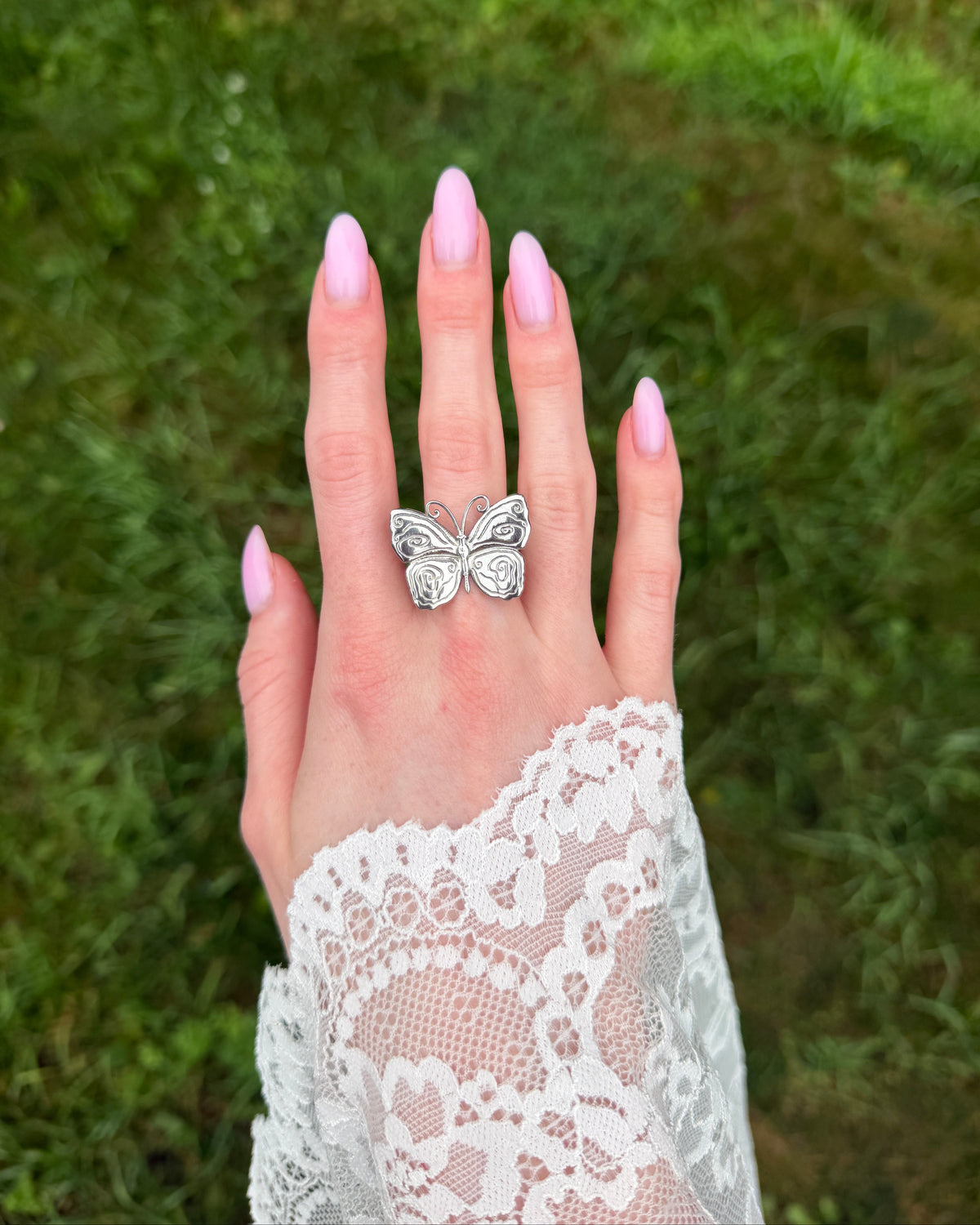 A hand with light pink manicured nails and a white lace sleeve, wearing a silver butterfly ring, set against a blurred green grass background.