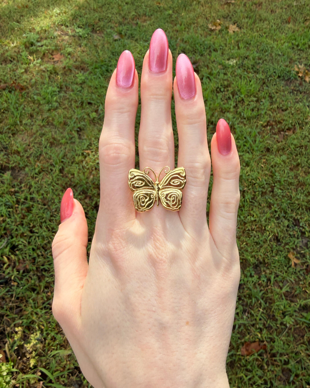A gold butterfly ring worn on a hand with pink manicured nails, set against a blurred green grass background.