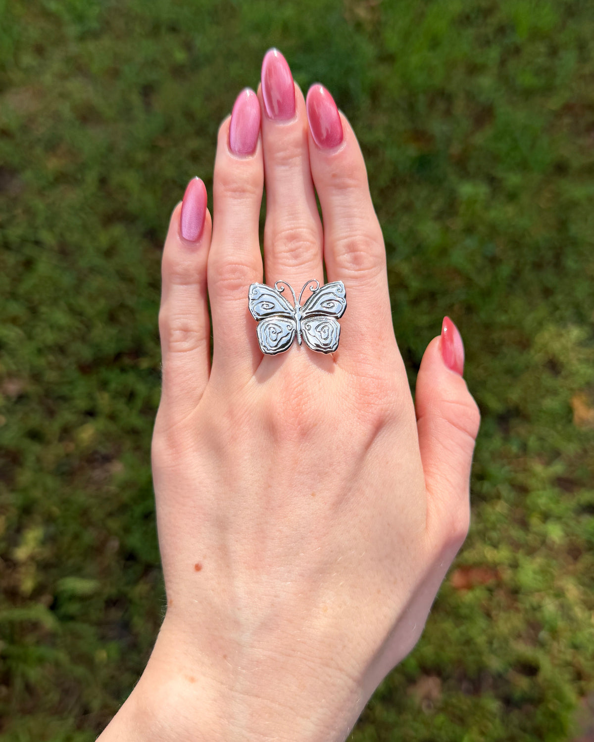 A silver butterfly ring worn on a hand with pink manicured nails, set against a blurred green grass background.