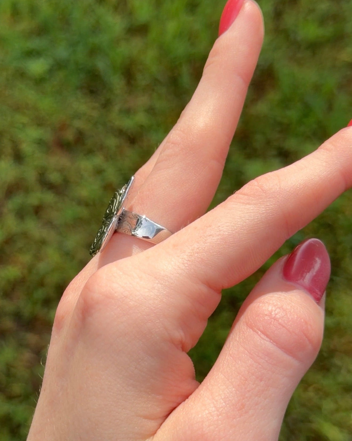 A close-up of a hand wearing a silver ring with a textured, organic-shaped band, set against a blurred green grass background.